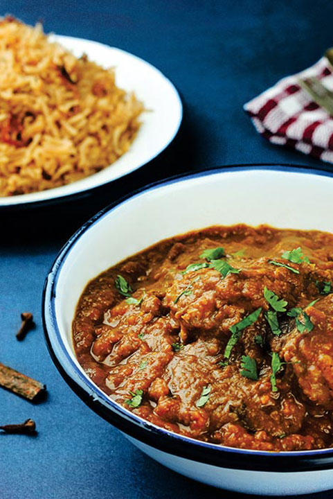 A bowl of rich chicken curry with herbs, served alongside a plate of seasoned rice and a small dish of onion relish, on a blue surface with a checkered napkin.