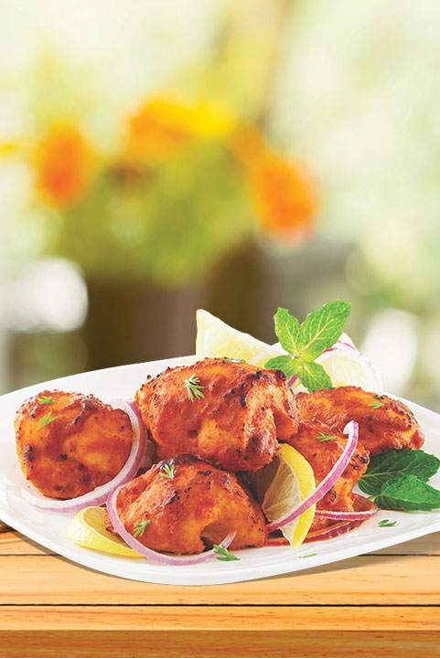 A white plate with chicken tikka pieces, garnished with lime wedges, red onions, and mint leaves, served on a wooden table with a blurred outdoor background.