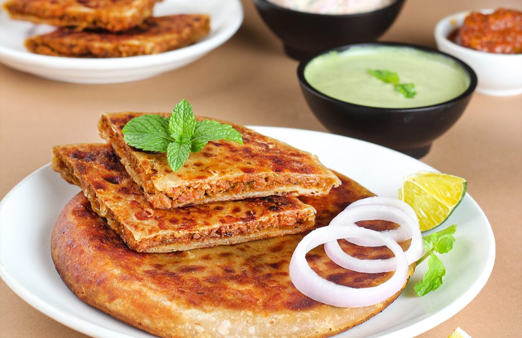 Golden chicken keema paratha served hot with sliced onions, lemon wedge, mint garnish and bowls of chutney and yoghurt dip
