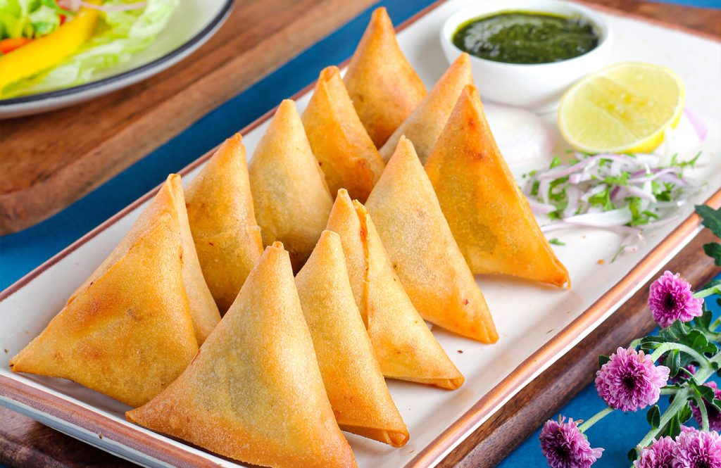 Crisp chicken keema samosas arranged on a tray with onion slices, lemon wedge and a small bowl of green chutney