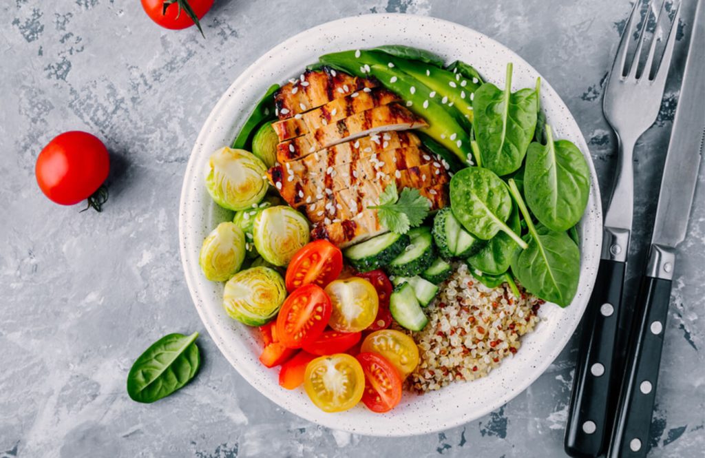 A bowl with grilled chicken, quinoa, spinach, Brussels sprouts, cucumber, and cherry tomatoes, garnished with sesame seeds, with a fork and knife on a grey surface.