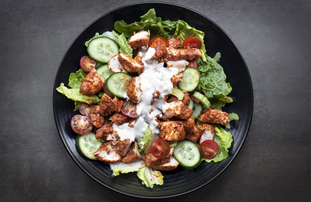 Black bowl with spicy chicken, lettuce, cucumber, tomatoes, and creamy dressing on a dark surface.