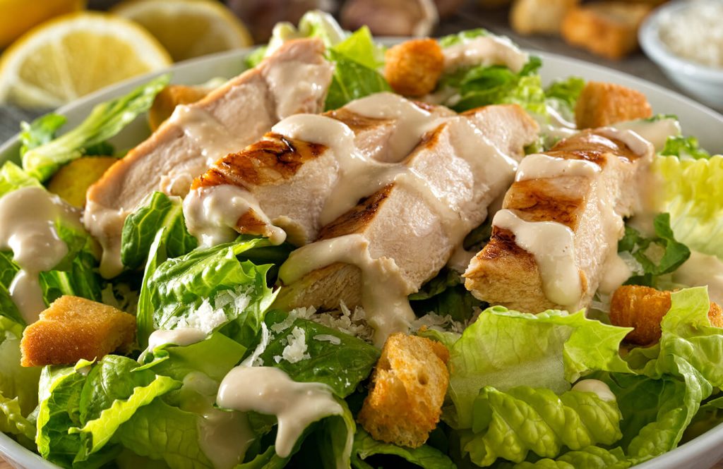 A bowl of Caesar salad with grilled chicken slices, croutons, romaine lettuce, creamy dressing, and grated Parmesan, with lemon wedges in the background.
