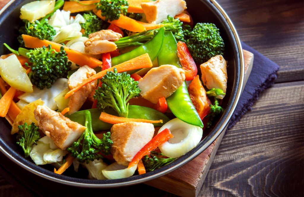 Vibrant chicken stir-fry with broccoli, carrots, bell peppers, snow peas, onions, and cabbage in a black wok on a wooden board.