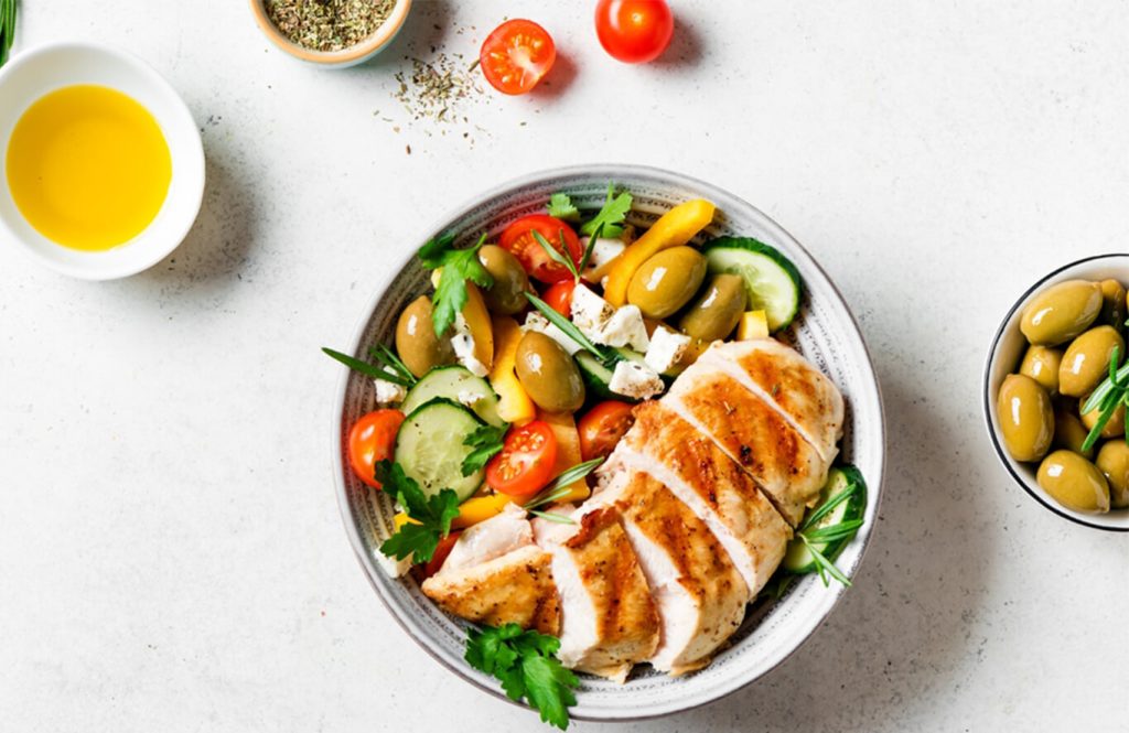 A grey bowl with grilled chicken breast, cucumber, tomatoes, olives, and feta cheese, served with olive oil and a side of olives on a white surface.