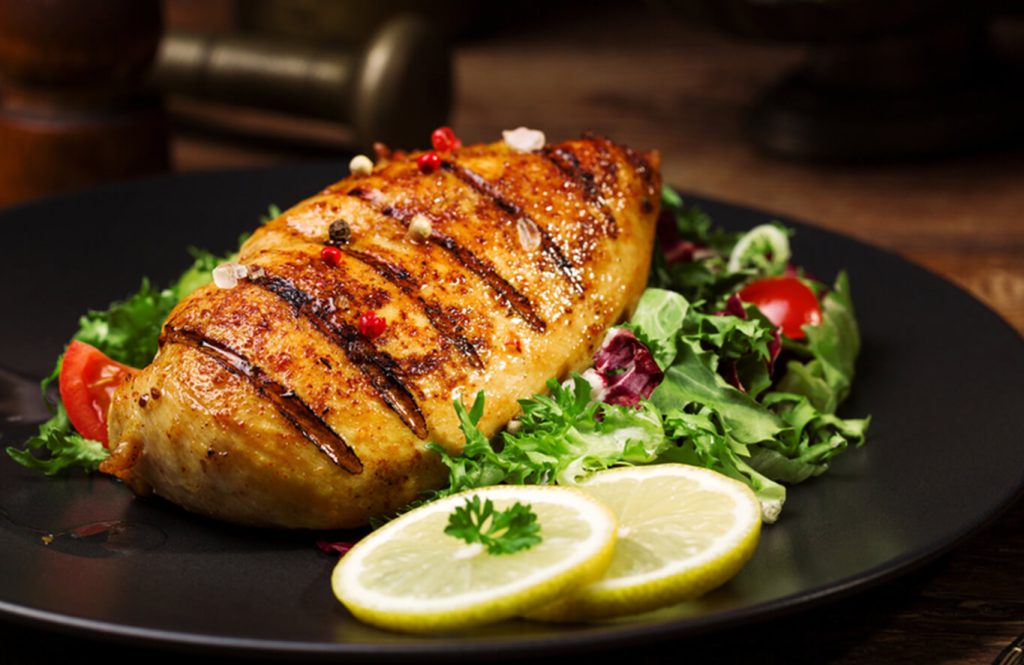 Grilled chicken breast with salad and lemon slices on black plate.