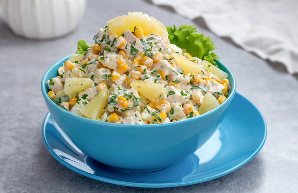 A bowl of salad with chicken, pineapple, corn, and parsley, garnished with lettuce leaves, served on a matching blue plate.