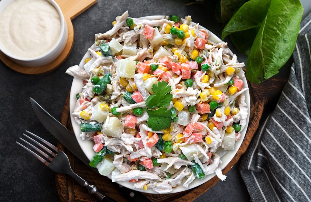 A white bowl of chicken salad with potatoes, peas, corn, diced carrots, and parsley, served with a side of creamy sauce and a fork and knife.