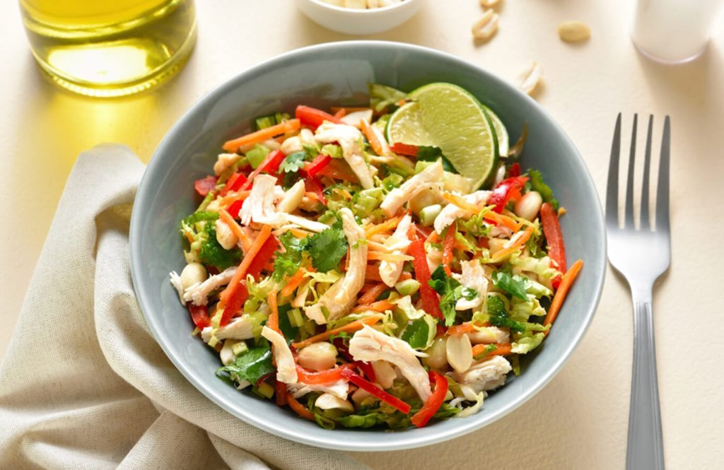 A blue bowl of fresh salad with shredded chicken, red bell peppers, carrots, cabbage, cilantro, and lime wedge, with a fork and scattered peanuts.