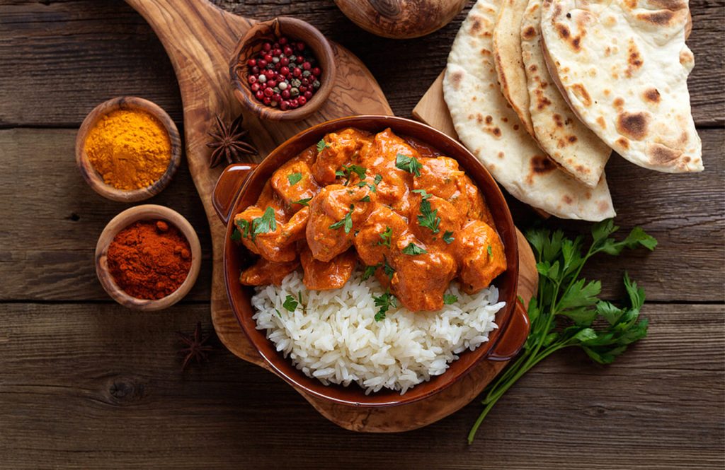 Bowl of creamy butter chicken over white rice, garnished with cilantro, with spices like turmeric, chili powder, black pepper, and naan bread on a wooden table.