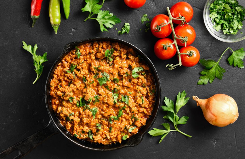 A pan of minced chicken curry simmering with onions, tomatoes, and spices, garnished with fresh coriander, served with rotis on a wooden table.