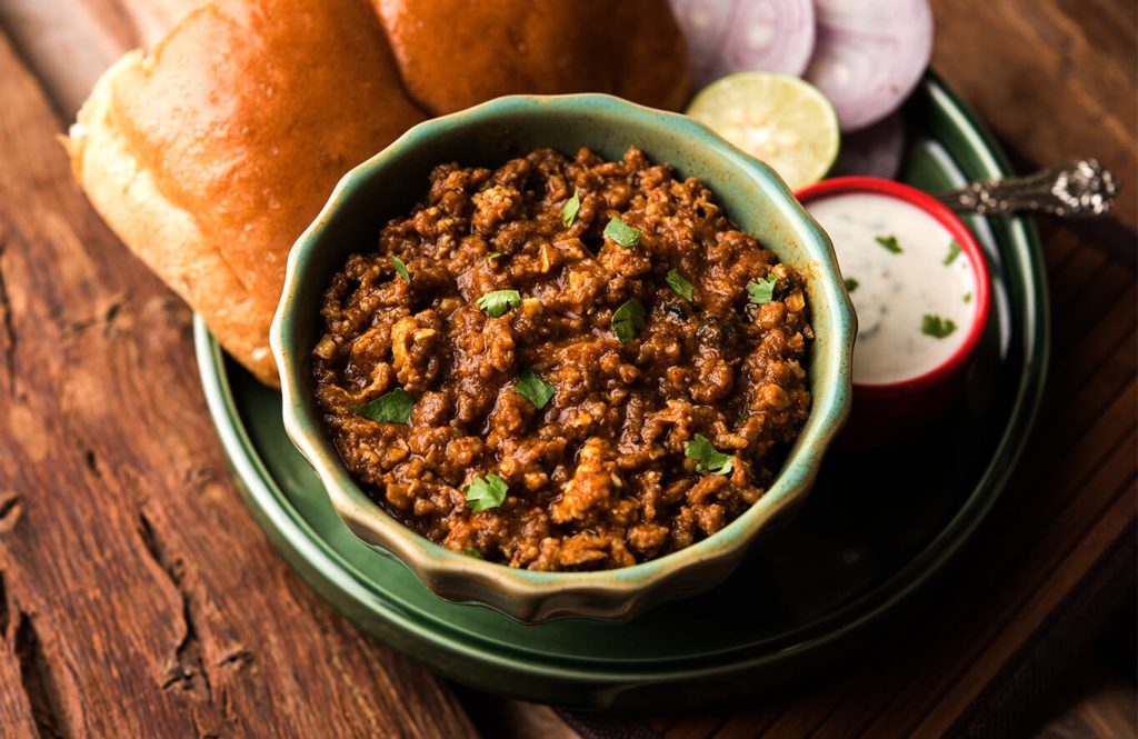Traditional chicken keema curry served with soft bread rolls, sliced onions, lemon wedge and cooling yoghurt dip on a rustic wooden table