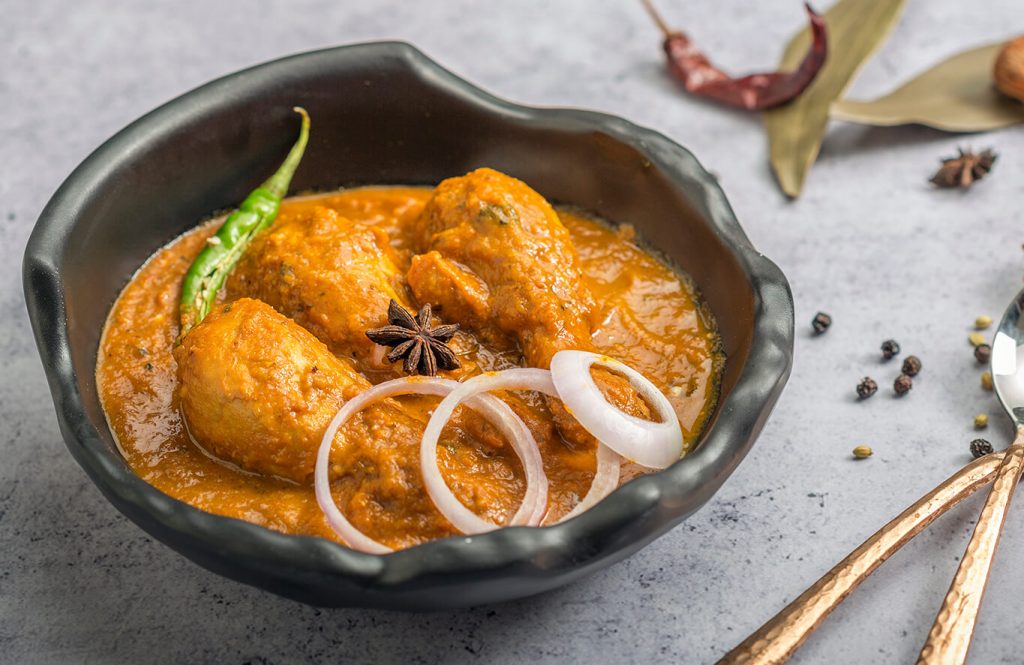 A bowl of rich curry with chicken pieces, garnished with green chili, onion rings, and star anise, served with spices and bay leaves on the side.