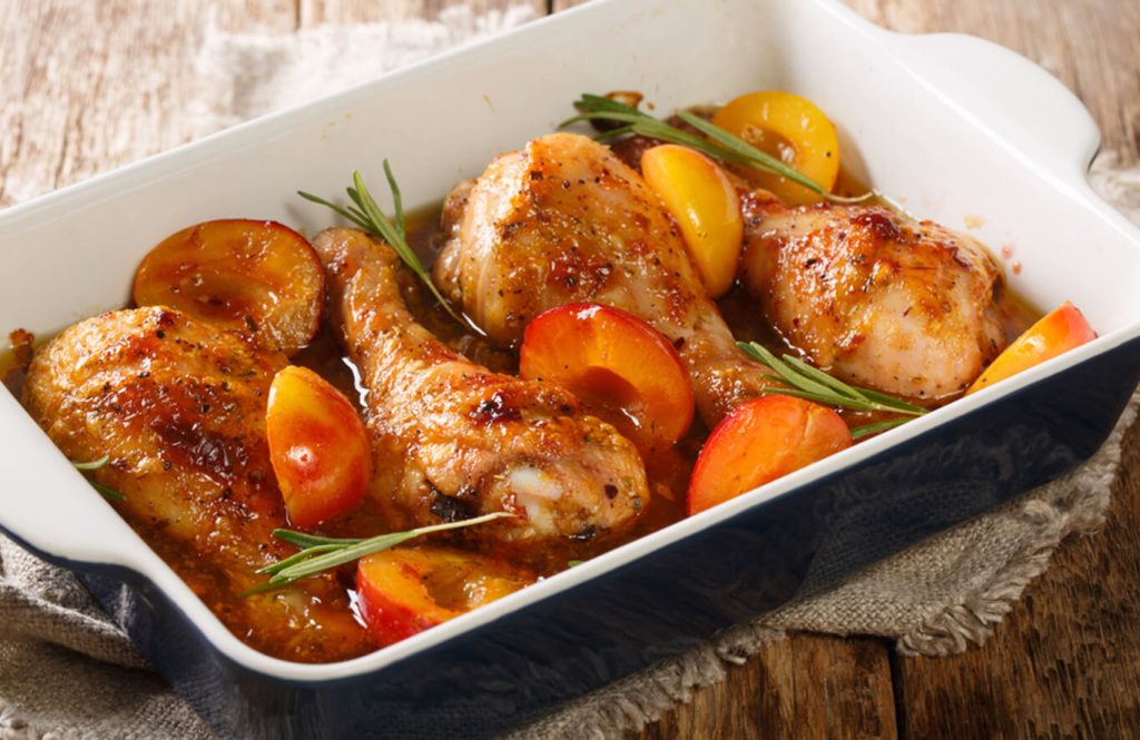 A white baking dish with roasted chicken drumsticks and apricot halves, garnished with rosemary, on a wooden surface with a cloth.