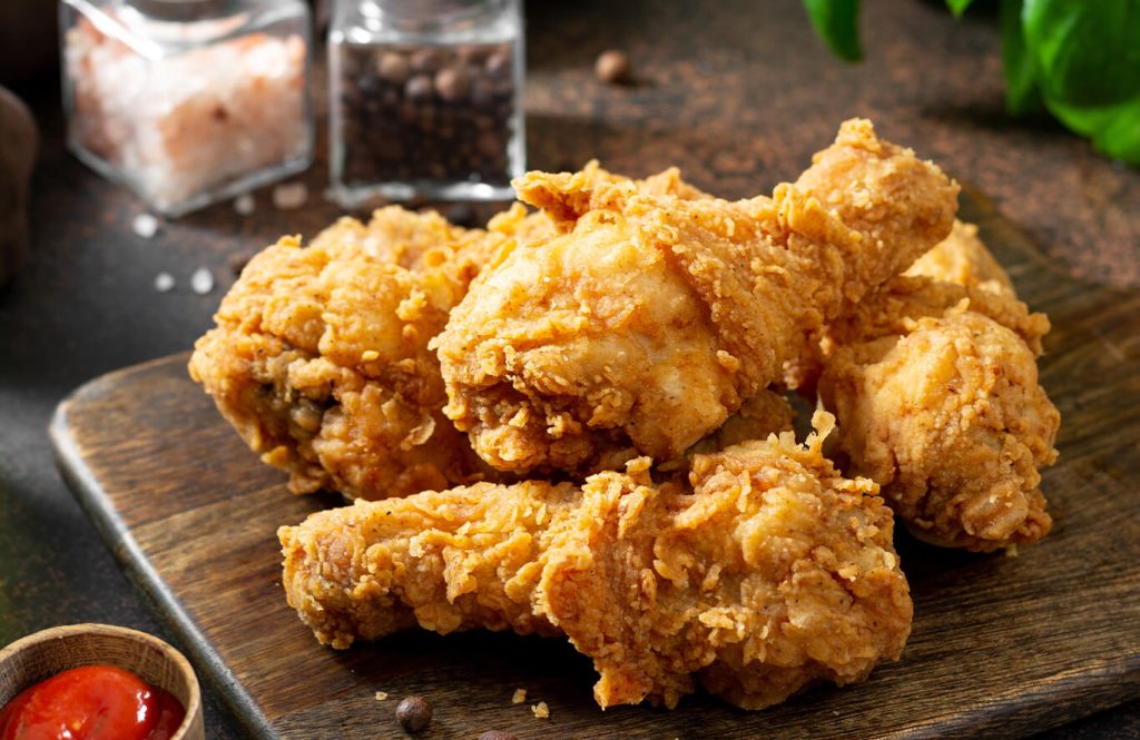 Crispy fried chicken drumsticks arranged on a wooden board, with sea salt, black pepper, and a small bowl of tomato ketchup.