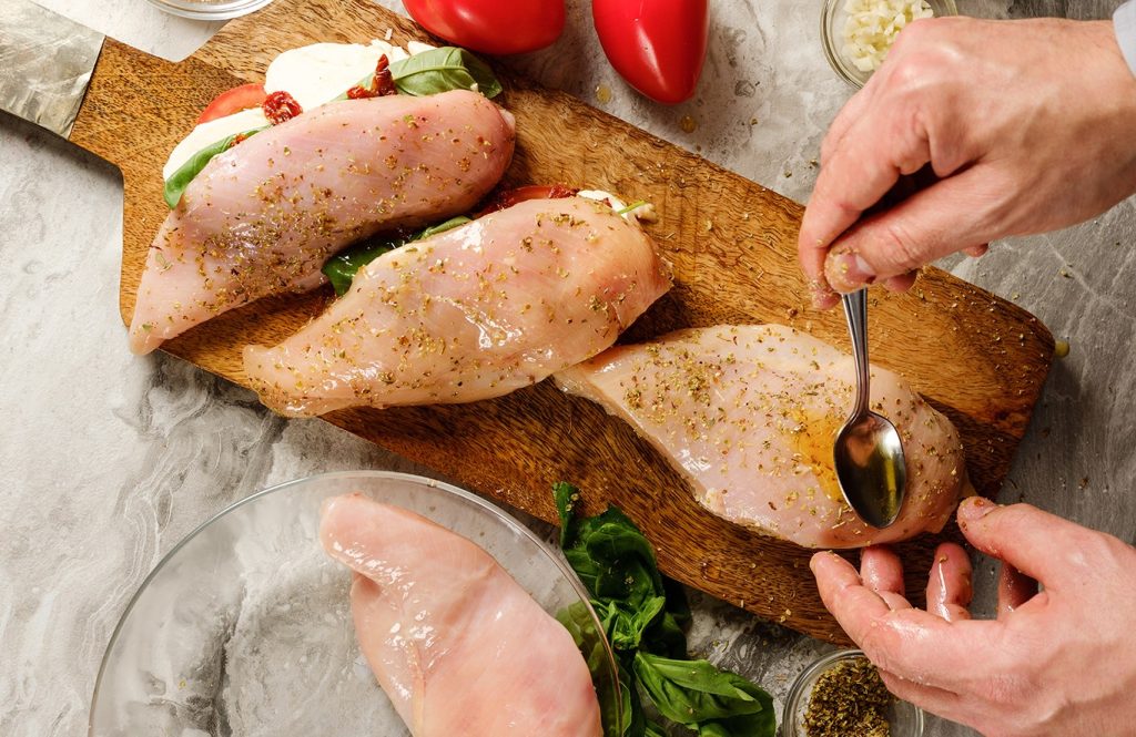 Hands stuffing raw chicken breasts with mozzarella, sun-dried tomatoes, and basil leaves on a wooden board, surrounded by red peppers, garlic, and herbs.