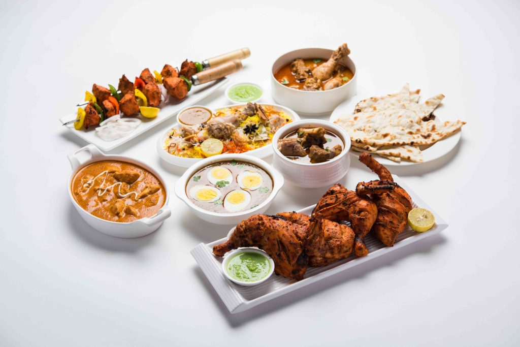 Assortment of Indian chicken dishes including butter chicken, chicken biryani, chicken tikka skewers, haleem, roasted chicken legs, egg curry, and naan bread on a white table.