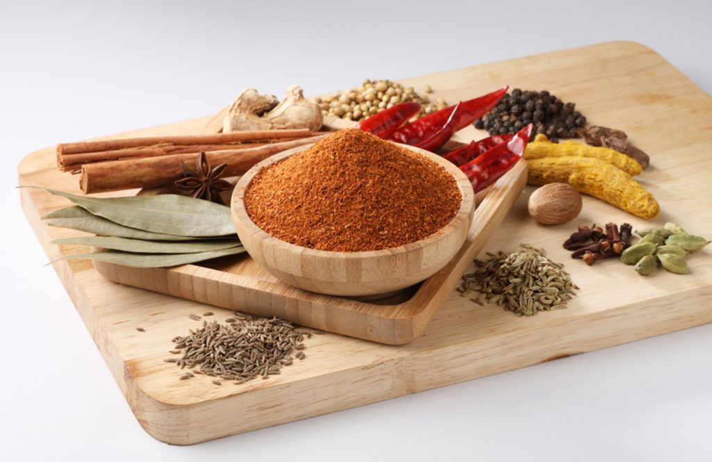 Malvani masala powder in a wooden bowl surrounded by whole spices such as cinnamon, cardamom, dried chillies, bay leaves, and cumin seeds on a wooden board.