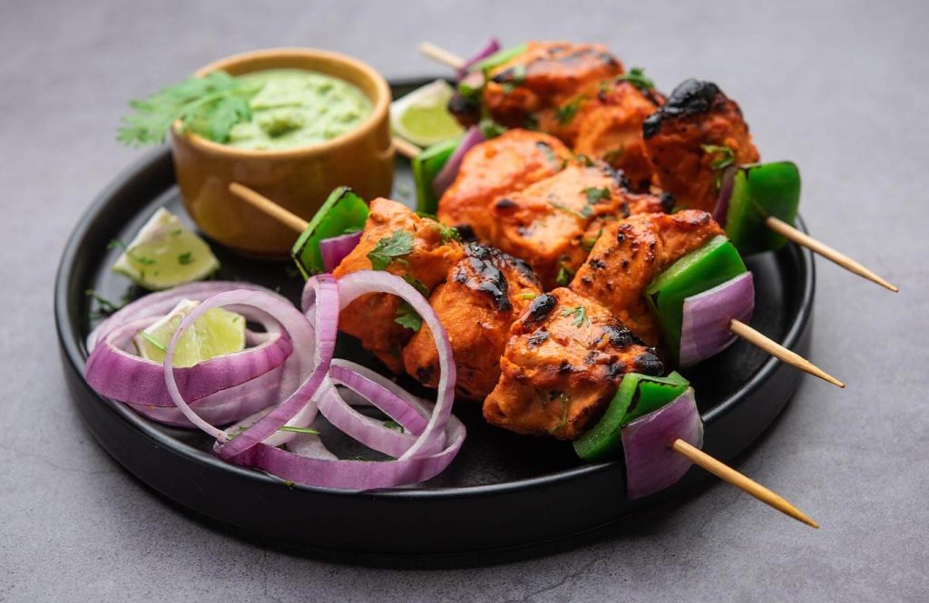 Grilled chicken tikka skewers with mint chutney on black plate.