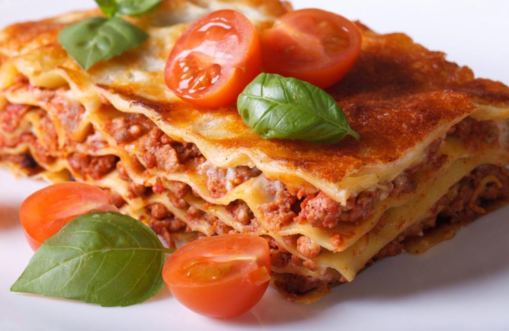Slice of spicy chicken keema lasagne layered with pasta, rich meat sauce, melted cheese, cherry tomatoes, and fresh basil leaves.