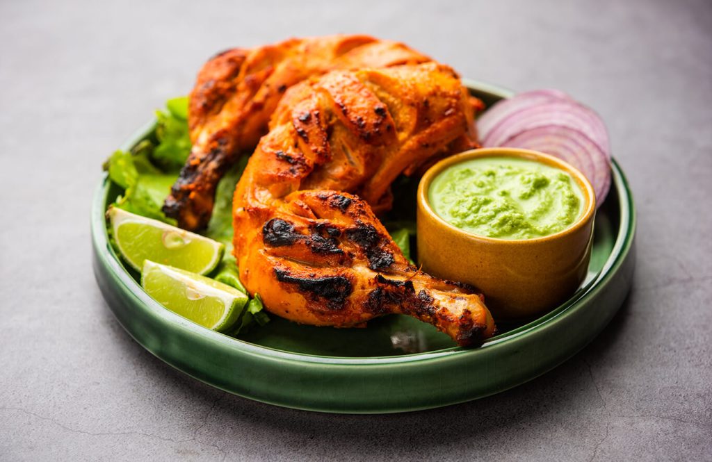Grilled tandoori chicken legs on a green plate with lettuce, lime wedges, green chutney dip, and onion rings on a gray surface.