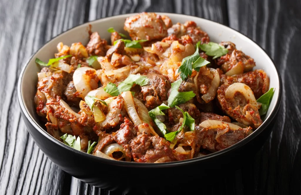 Pan-cooked chicken liver pieces with cumin-spiced onions, garnished with parsley, served in a clay bowl.