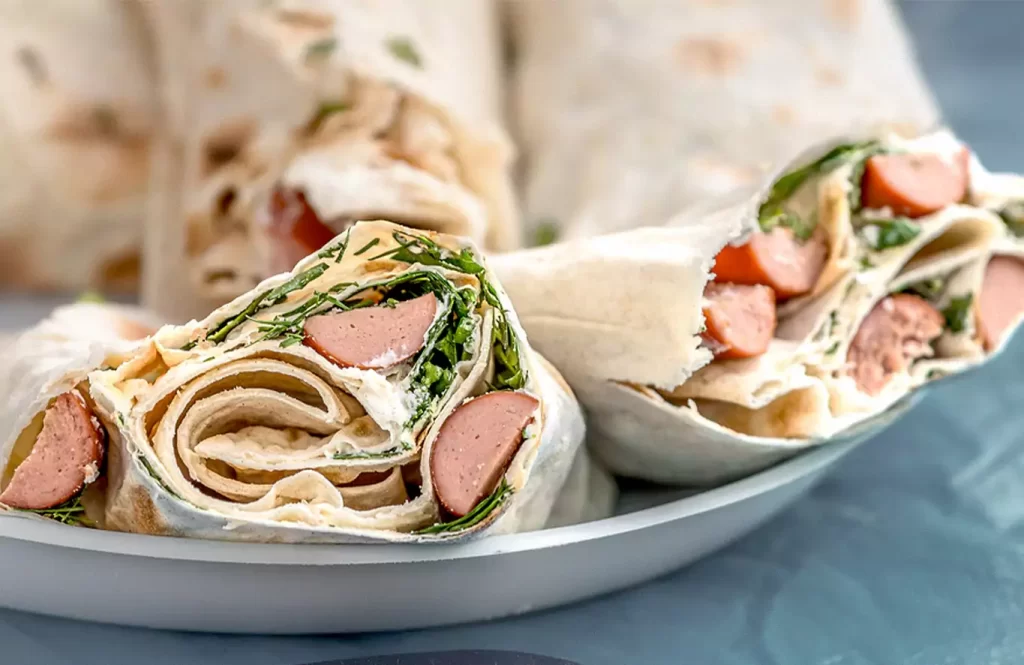 Sliced spinach tortilla roll with chicken sausages and sautéed spinach, spread with cream cheese, on a wooden platter.