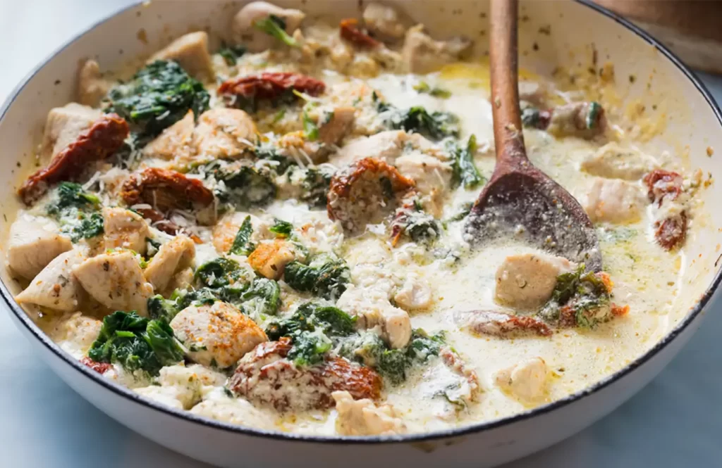A skillet with creamy chicken and spinach dish, featuring chicken cubes, wilted spinach, and a wooden spoon, served on a light blue surface.