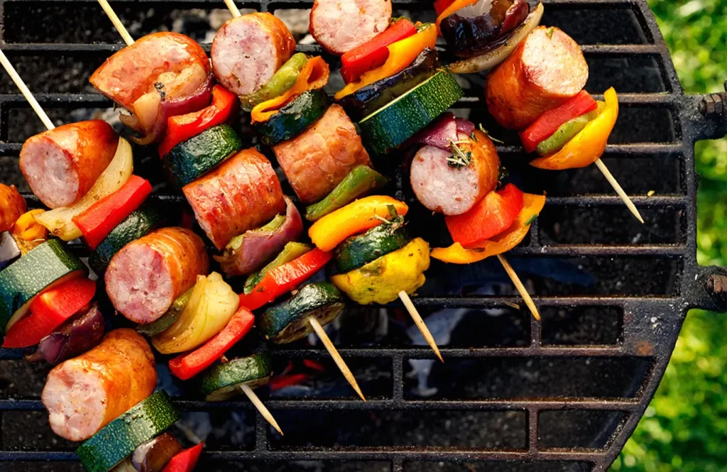 Grilled skewers of chicken cocktail sausages, cherry tomatoes, and bell peppers on a barbecue grill.