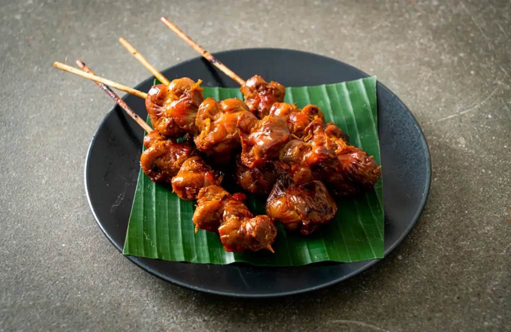 Grilled chicken gizzard skewers on wooden sticks, arranged on banana leaves in a black plate, with glossy orange marinade.