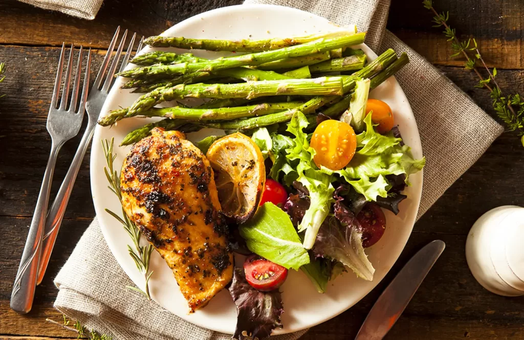 Grilled chicken breast with tender asparagus spears in a light broth sauce, served on a white plate.