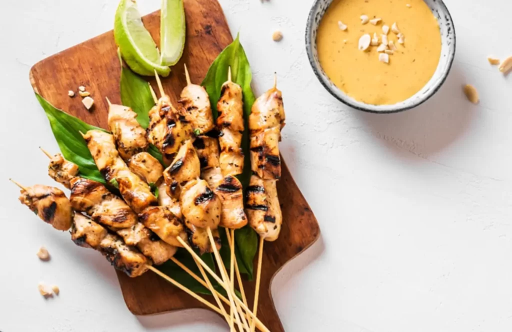 Grilled chicken satay skewers served on a wooden board with lime wedges and a bowl of peanut sauce
