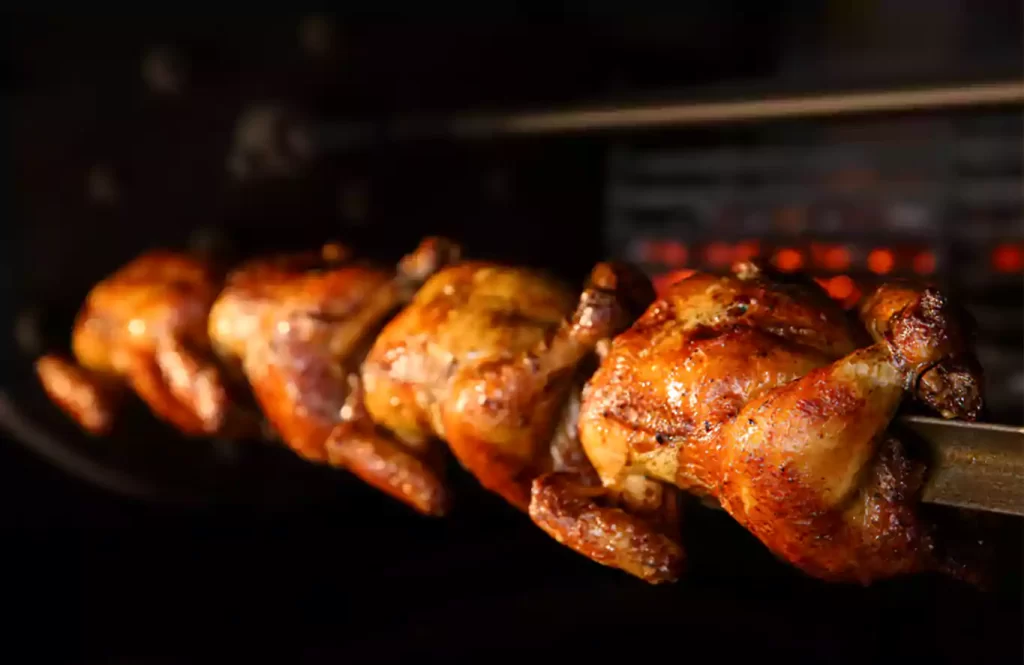Whole chicken roasting on a spit over a charcoal fire, with herbs and spices visible, set against a rustic outdoor backdrop.
