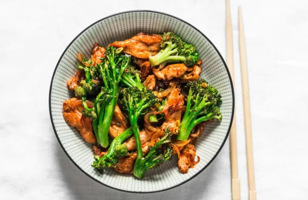 A bowl of stir-fried chicken and broccoli with a glossy sauce, served with chopsticks on a white tablecloth.