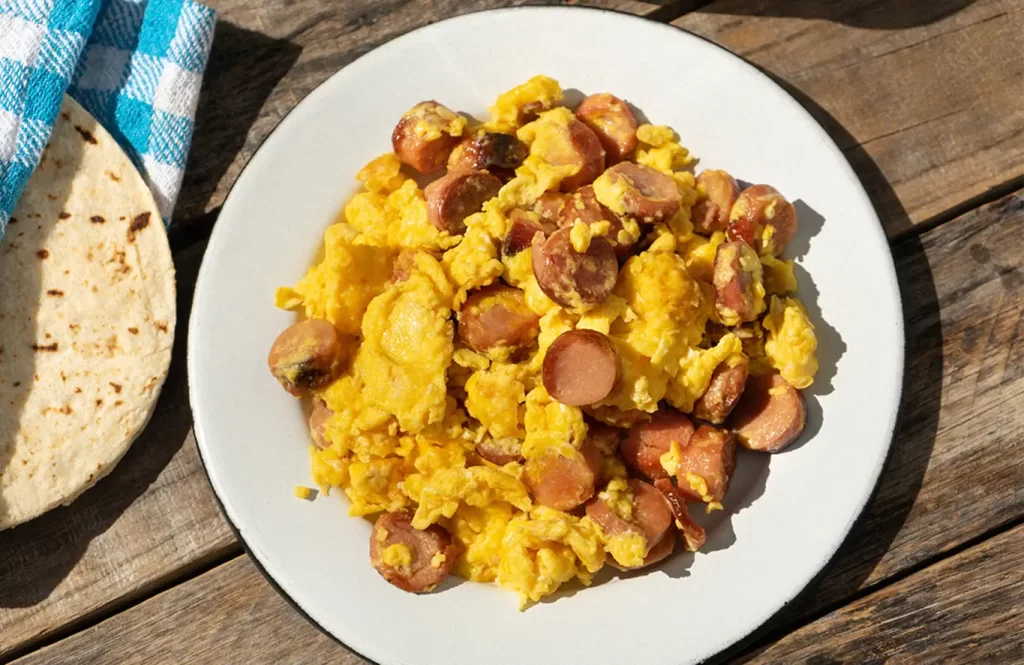 Scrambled eggs with sliced chicken frankfurters and diced potatoes, garnished with coriander, served with a tortilla on a wooden table.