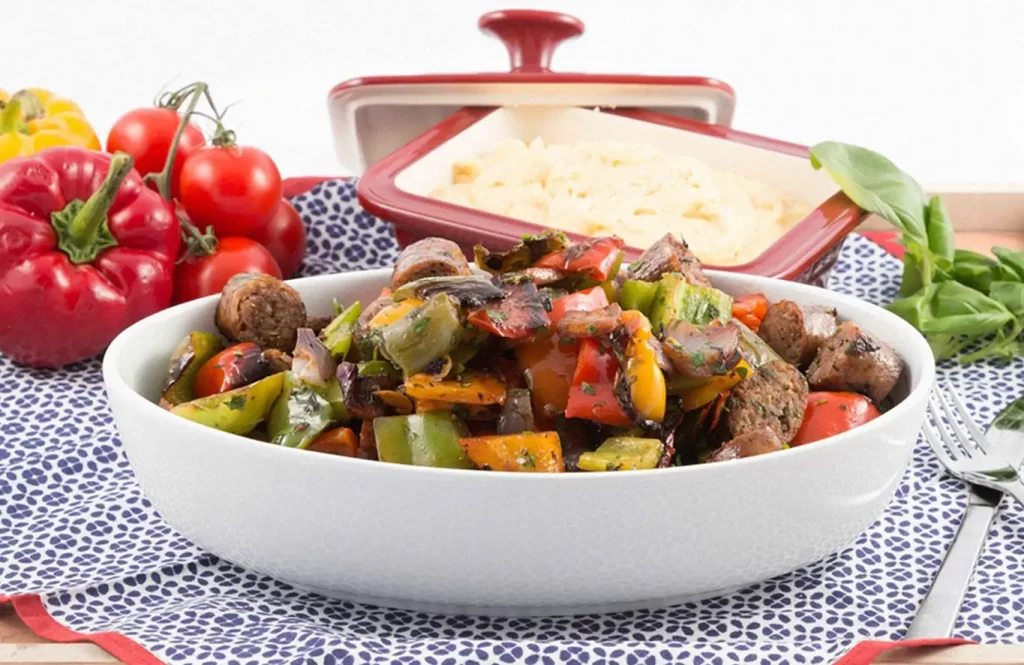 Spicy chicken sausage stir-fry with bell peppers and onions, served with jeera rice, alongside fresh vegetables and a red casserole dish.