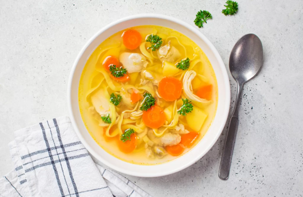 A bowl of homemade chicken broth with carrots, noodles, and parsley.