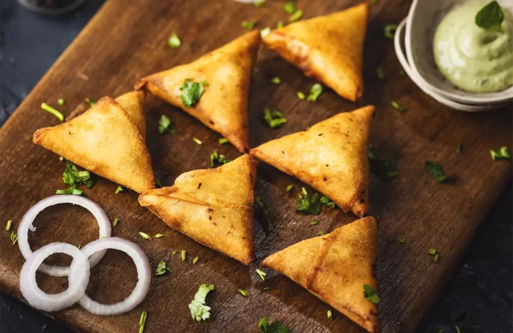 Golden-brown chicken samosas served with mint chutney and onion rings on a wooden board.