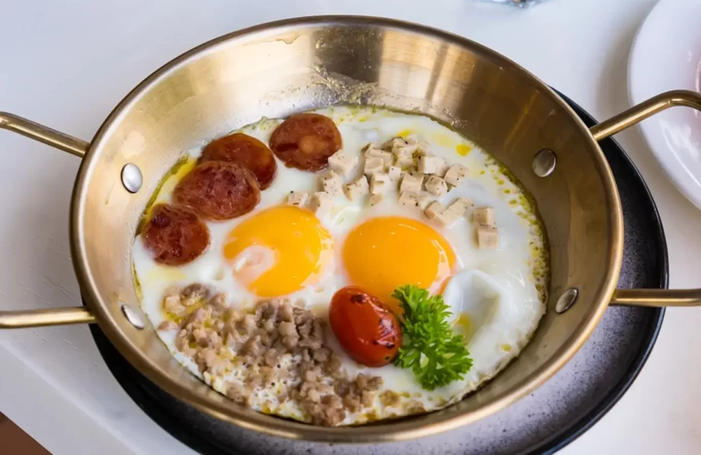 Sunny-side-up eggs served in a brass skillet with chicken sausages, ground chicken, cubed chicken, cherry tomato, and parsley garnish.