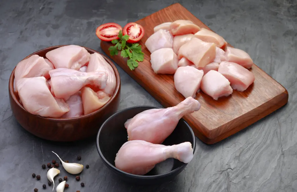 Fresh raw chicken portions including boneless breast, lollypops, skinless drumsticks, boneless leg, skinless legs and liver displayed on wooden board with tomatoes, garlic, peppercorns and herbs.