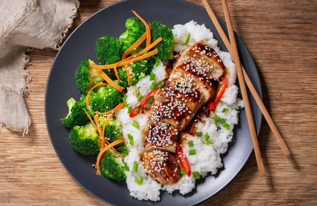 Sliced chicken teriyaki with a glossy soy glaze served over steamed rice with sesame seeds and spring onions on a dark plate