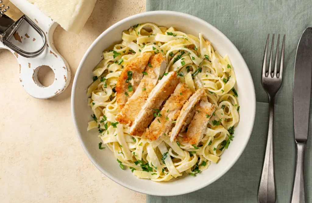Creamy chicken pasta with sliced breaded chicken on fettuccine tossed in a rich parmesan sauce, garnished with fresh parsley in a white bowl on a green cloth.