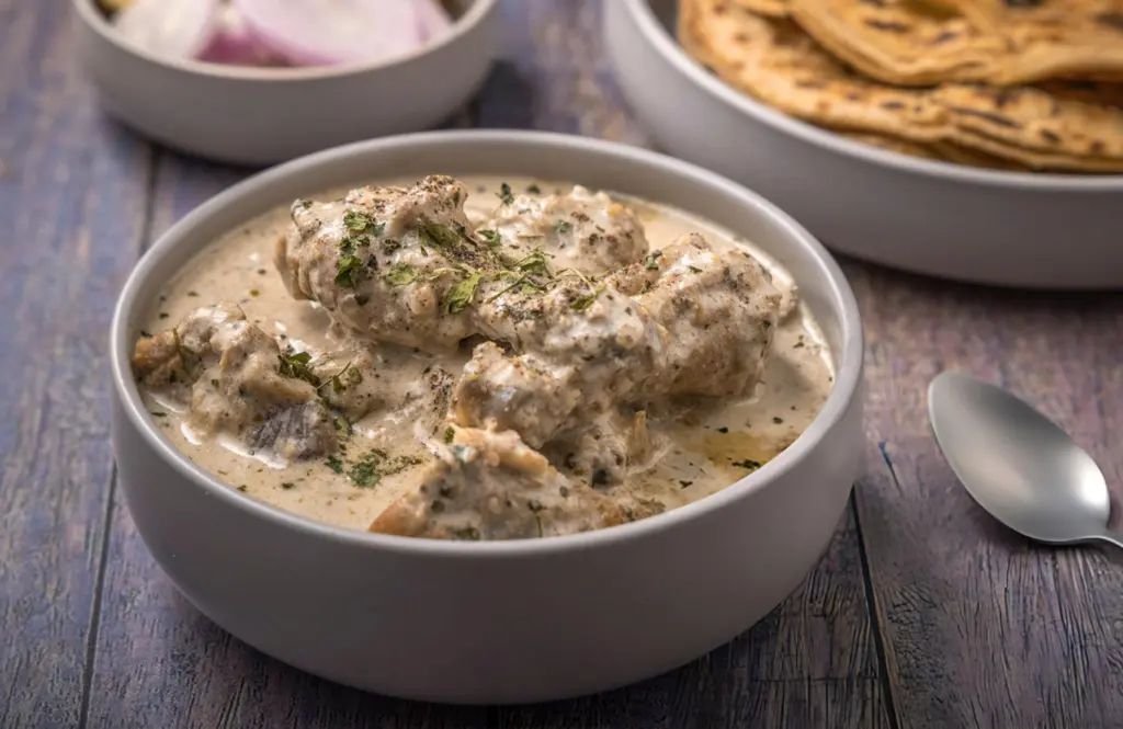 Creamy Mughlai chicken korma in a white bowl with visible whole spices served with naan on a wooden surface