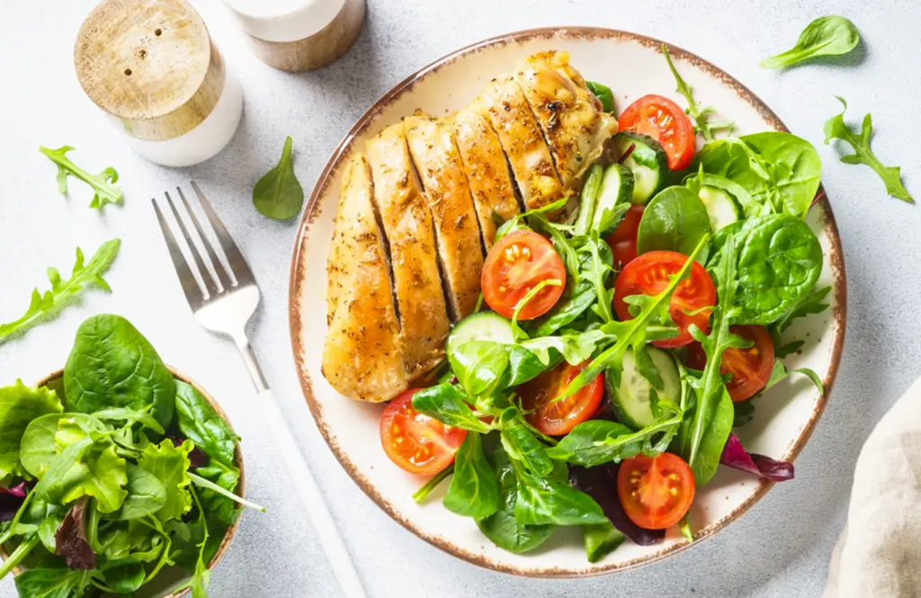 Sliced grilled chicken breast on a plate with fresh mixed greens, tomatoes and cucumber for a healthy salad
