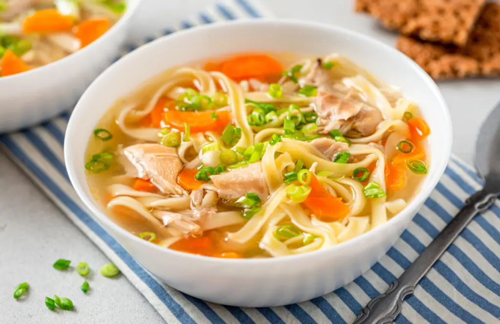Bowl of homemade chicken noodle soup with tender shredded chicken, wide egg noodles, sliced carrots, and chopped green onions in a clear broth, served on a blue striped cloth with crackers in the background