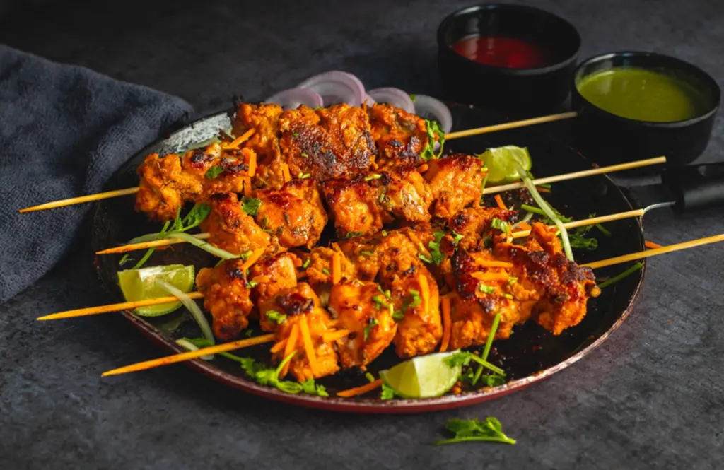 Grilled chicken tikka skewers on a dark plate with charred pieces of marinated chicken, garnished with fresh coriander, carrot juliennes, onion rings, lime wedges, and served with red and green chutneys