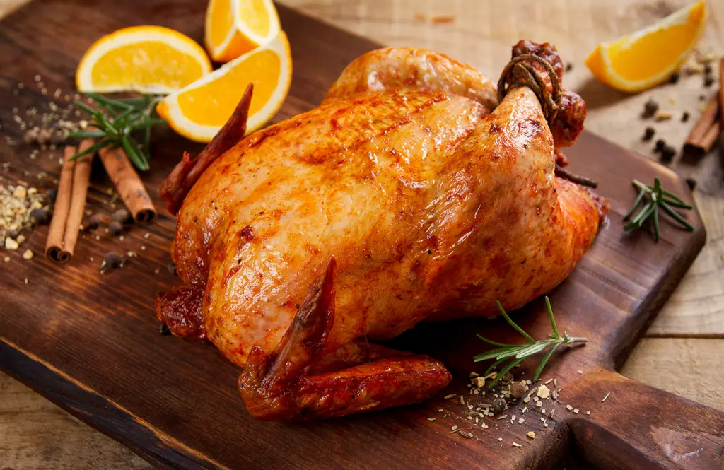 Roasted whole chicken with golden crispy skin on a wooden board, garnished with orange slices, rosemary sprigs, cinnamon sticks, peppercorns, and spices, ready for carving
