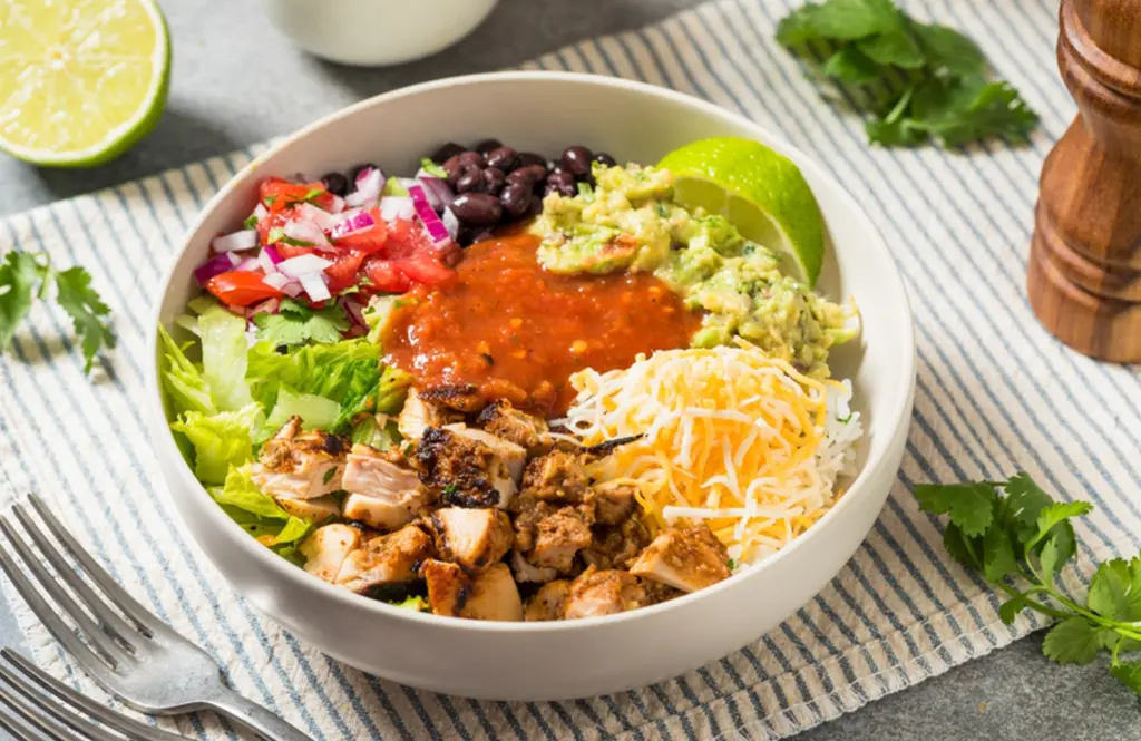 Chicken burrito bowl with grilled chicken, rice, black beans, salsa, guacamole, cheese and lime in a white bowl