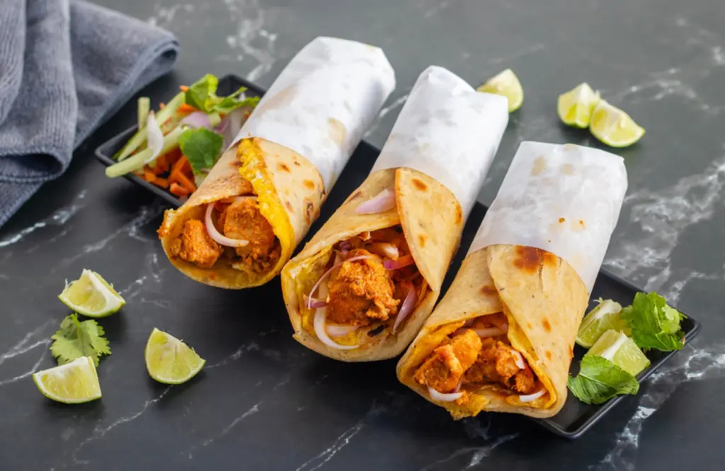 Chicken paratha rolls wrapped in parchment paper with marinated chicken, onions and vegetables on a black plate