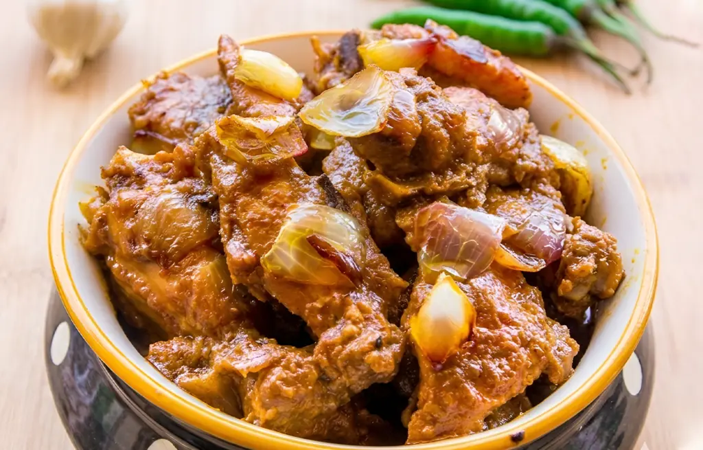 Delhi-style Chicken Do Pyaza made with Zorabian Chicken Curry Cut in a thick spiced masala with caramelised onion petals, served in a dark ceramic bowl with green chillies and garlic